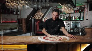 Experienced pizzaiolo in black chef's uniform adding grated cheese on round pizza base. Male chef cooking italian meal according to traditional recipe.