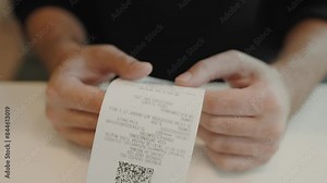 A person examines a detailed receipt with a QR code. The focus is on the hands and the printed paper, capturing a moment of financial transaction or review. Man Reviewing Electronic Receipt