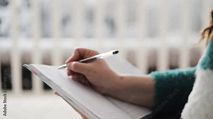 Young woman spending free time in bedroom, sitting on bed cover, thinking of plans for week and writing them in notebook that is on her knees.