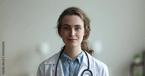 Pretty young medical specialist, hospital intern, doctor woman head shot portrait. Serious practitioner in white coat looking at camera, getting happy, cheerful, smiling