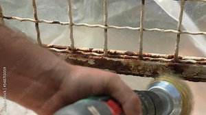 Electric grinder with round metal brush nozzle rotating at an angle and removing old rusted paint from metal surface, top view shot of workman hands holding grinder machine and removing rust remains