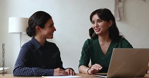 Two attractive friendly women colleagues talking in office at workplace desk, share thoughts, ideas, opinions enjoy break and pleasant informal conversation during workday, having successful team-work