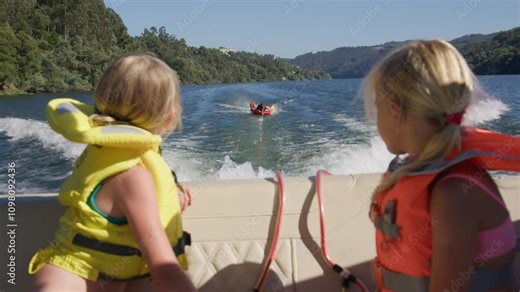 Two girls in life vests sitting in a boat watching a person on an inflatable tube being towed behind on a scenic river