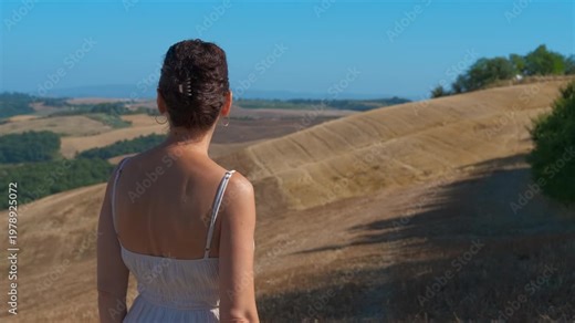 Elegant woman enjoying sunny tuscan hills landscape. Beautiful mature woman in a summer dress and sunglasses standing and admiring the panoramic view of the golden rolling hills under a blue sky