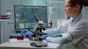 Biochemistry doctor analyzing vacutainer with blood while having microscope on desk and using computer in professional medical laboratory. Specialist woman doing analysis experiment