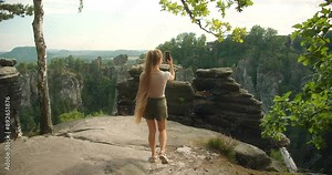 Woman walks on a cliff edge taking a selfie with the impressive Bastei rock formations and a picturesque landscape in the background