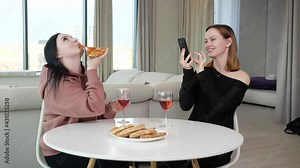 two friends have fun drinking wine, eating pizza and taking pictures on the phone while sitting at home at the table. High quality 4k footage