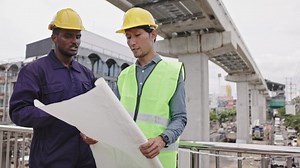 Structural engineers and construction workers are holding blueprints inspecting structure. sky train track construction in city. Blue collar wears a safety hardhat while working on construction site.