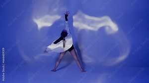 Young woman wearing a top, shorts and a shirt performing contemporary dance under the glare of light in the studio. Neon blue color scheme, shadowed background. Full length.
