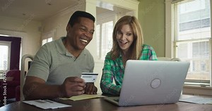Happy senior couple booking trip or making purchase online with credit card using laptop computer. African American and Caucasian husband and wife paying off debt. Slow motion 4k handheld
