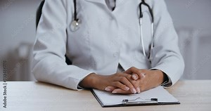 Close up part of female body of unknown woman with black skin, doctor or nurse sitting at table, wears uniform, put her hands on notepad, documents, paper, folded fingers in lock and breathes deeply