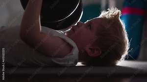 Strong little boy tries to lift heavy barbell lying on floor imitating adult in gym. Motivated kid enjoys training with weight to pump up arm muscles slow motion
