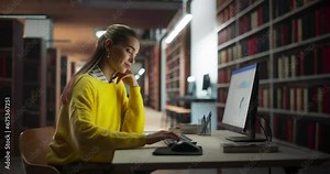 Young Female Student in Yellow Jumper Working on Her Master's Thesis on a Desktop Computer. Smart Woman Learning the Lecture Online, Looking Through a Presentation in a Library with Book Shelves