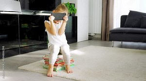 Little girl sits on stack of children's books and watches cartoons on smartphone