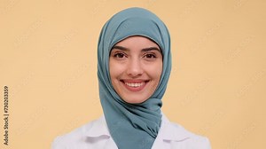 Close up portrait of a Muslim woman wearing on hijab and lab coat nurse smiling at camera over beige background. Positive medical service, cheerful hospital staff.