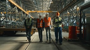 Four heavy industry engineers in safety uniform and hard hats discuss work process walking at manufacturing factory. Mature supervisor holds laptop, talks to diverse workers in warehouse. Slow motion.
