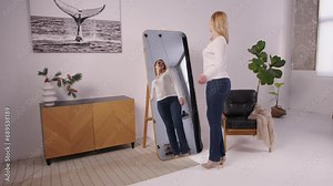 Joyful attractive middle aged woman with perfect body in stylish clothes and high heels looking at reflection in mirror, admiring appearance and shapes after successful weight loss at home.