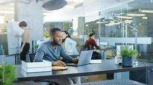 Busy International Office, African-American Man Working at His Desk on a Laptop, He Waves Hello to the Passing Colleague, in the Background Businesswomen Discuss Relevant Data.