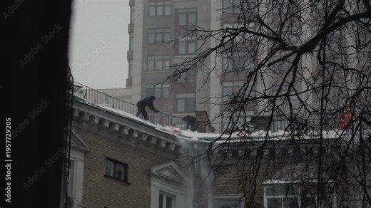 Rope access snow shoveling from roof of building. Man uses a shovel to clean a house during a blizzard. Snowfall in winter in Russia. Male worker janitor throws or drop ice, frost. Apartment building