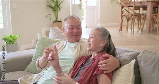 Mature Asian couple reacting to man smiling on sofa in living room, smile-to-laugh with glasses