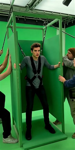 This behind-the-scenes footage reveals how terrifying skyscraper free-fall scenes are safely filmed. The actor performs inside a small green elevator frame made of metal bars and foam panels, secured by a safety harness. Crew members pull thick cables downward in short, uneven jerks to simulate falling motion while the director loudly counts down each reaction cue. The camera remains handheld, slightly tilting and refocusing mid-take to capture realistic panic. Green padding is visible below, wi