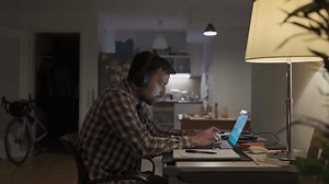 University student learning by laptop with notebook and pen. Online class, E-Learning. Guy wearing headphones typing on keyboard of notebook and making notations in notebook late at night at home.