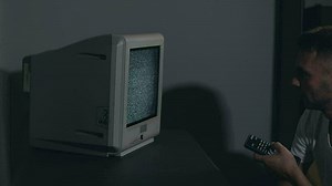 Broken Television or TV Propaganda. Mind Control - Zombified Man in White T-shirt Switches Channels on the Old TV in Dark Room. TV Signal Search. Distortion on the Screen. Medium Close-Up, Film Grain