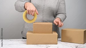 woman in a gray sweater holds a roll of duct tape and packs brown cardboard boxes on a white table, behind a stack of boxes. Moving concept