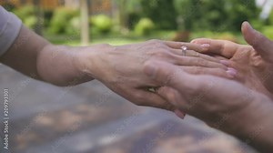 Close-up senior female hand getting engagment ring on her finger from elderly boyfriend. Senior male making marriage proposal to fiancee outdoors. Female looking and admiring her diamond ring