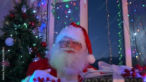 Angry bad Santa shows obscene gestures with his hands fingers sitting next to pile of New Year's gifts and a Christmas tree holiday background.