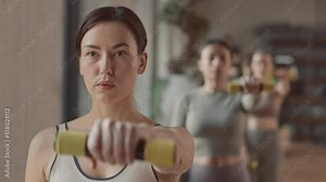 Chest up slowmo of young female trainer doing alternating dumbbell front raise and group of women repeating after her, having indoor workout in fitness studio