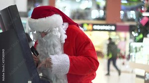 Santa Claus inserting credit card into bancomat entering a pin to receive money. Santa Claus withdrawing money from cash dispenser in a large shopping center