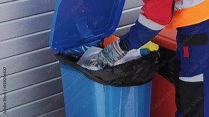 Waste management worker collects recyclables from a blue trash bin in an urban setting on a sunny day in September