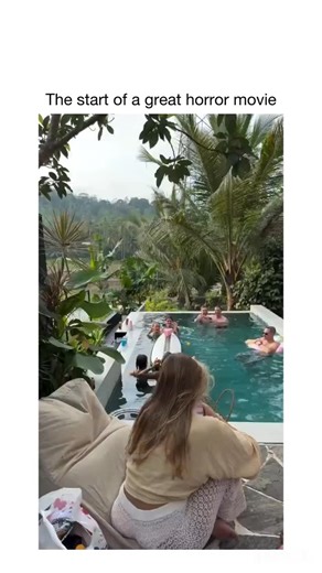 LaughOrbit on Instagram: "Video footage of an outdoor infinity pool set amid lush tropical vegetation and distant hills under an overcast sky. A group of people, including men and women in swimwear, relax and play in the turquoise water using surfboards, inflatable rings, and drinks. A woman wearing a light top and white pants sits on a beige lounge chair in the foreground, facing the pool. The camera pans downward through palm fronds and greenery, revealing a dark-skinned individual partially o