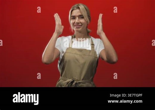 Woman wearing khaki apron frames her chin with right hand, lifts left arm and smiles showing teeth on a red studio set; playful joy Stock Video Footage - Alamy