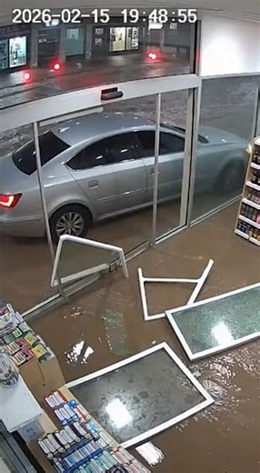 CCTV security camera outside a local store faces the front windows as heavy rain pounds outside. In the opening seconds, floodwater rushes across the street, headlights reflecting off fast moving water. A car suddenly hydroplanes, losing all control, and slides sideways with the current straight toward the storefront. The vehicle slams through the glass entrance as water pours in behind it, knocking over displays and sending debris skidding across the floor. Alarms blare while muddy water spread