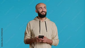 Arab person typing text messages on smartphone app, using mobile phone internet to check social media. Middle eastern guy browsing online websites in studio, blue backdrop. Camera 1.