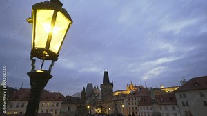 Illuminated Charles Bridge gas streetlight lantern against square with tourists admiring ancient architectural buildings with large spires under cloudy sky in twilight