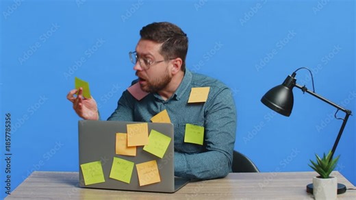 Exhausted man sitting at table with pasted sticker notes use laptop computer having concentration problem creative crisis. Nervous adult guy confused by big routine work loading on blue background