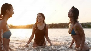 Happy wet girls in bikini run into sea and play splashing water to each other on sunset. Cheerful female friends have fun making splashes in pond. Young women go to swim, bathe in lake. Slow motion.
