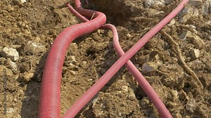 CLOSE UP: Corrugated plastic utility tubing lies inside a trench dug into the soil of a road under construction. Wide red plastic telecommunication conduits run along a newly dug up shallow ditch.