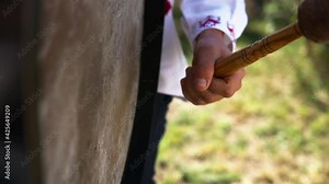 A caucasian man in an embroidered shirt plays on a big drum made of leather stretched on a resonator and beats it with a massive mallet with a soft tip and hear strong deaf and low sound