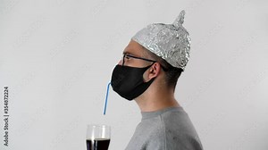 Man in a foil hat and a protective medical mask drinks beer through a tube from a glass. Portrait of a man drinking carbonated drink. 5G tower radiation protection. Quarantine self-isolation drinks