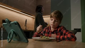 Child enjoys a meal while watching tablet computer. Mom intervenes, promoting healthy habits. Parenting and lifestyle content