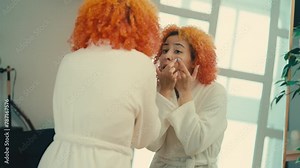 Young woman squeezing a pimple in front of mirror, face skin imperfections