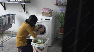 happy asian woman in front of the washing machine doing some laundry loading clothes inside