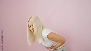 Painful bellyache, young blonde woman, doubled over in stomach pain, ill and suffering from abdominal cramps, stands isolated on a pink background