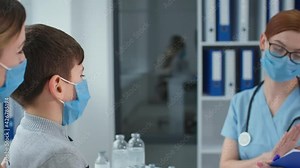 pediatrician in hospital, masked female doctor in glasses measures the temperature of boy patient with non-contact thermometer