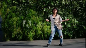 woman performs slow-motion dance, moving gracefully in outdoor setting.Dressed in casual attire female is immersed in rhythm,showcasing fluid and expressive movements against backdrop of lush greenery