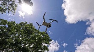 A drone rises through a clear sky, framed by trees and sunlight, capturing dynamic aerial movement
